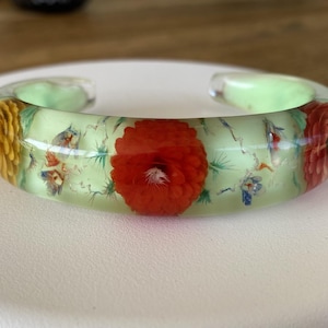 Peut inclure: Un bracelet jonc en résine vert clair translucide incrusté de fleurs séchées aux teintes orange vif, jaune et rouge profond. Ce bracelet ouvert présente des détails floraux préservés encapsulés dans une matière lisse et polie.