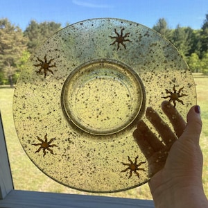 May include: A round, translucent yellow glass plate featuring a central circular indentation. The surface is embedded with numerous small gold star-shaped glitter pieces and four distinct dark brown sun-shaped motifs arranged symmetrically.