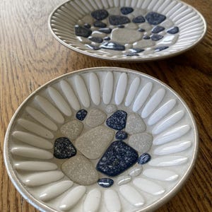 May include: Two round ceramic dishes featuring a decorative mosaic pattern. The rims are lined with elongated white petals, while the centers contain a mix of smooth, rounded beige and dark blue speckled stones embedded in a light base.