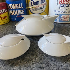 Puede incluir: Un juego de té de cerámica blanca que consta de una tetera con asa de metal, un azucarero y una jarra de leche. Cada pieza presenta una base circular de perfil bajo y un pico o tapa puntiaguda, creando una estética suave y redondeada.
