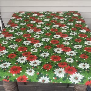 May include: A rectangular tablecloth featuring a vibrant green background covered in a dense, repeating pattern of red and white poinsettia flowers, green pine needles, and small white berries, draped over a table with dark metal chairs.