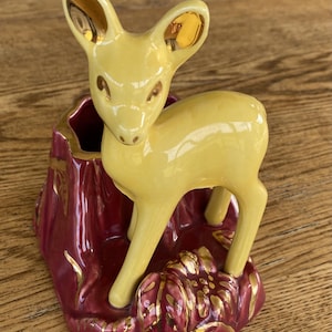 May include: A ceramic planter featuring a pale yellow fawn standing next to a dark red tree stump. The fawn has gold-painted accents on its ears and eyes, while the base is decorated with dark red leaves highlighted with gold paint details.