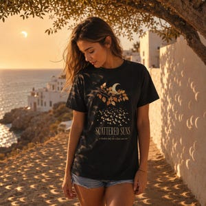 Puede incluir: Una camiseta negra de manga corta con un estampado gráfico de una luna creciente entre hojas de tonos dorados, con el texto "SCATTERED SUNS" y "a shadow only the eclipse can cast" impreso en una fuente serif clara debajo.