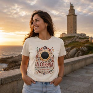 May include: A cream-coloured short-sleeve t-shirt featuring a graphic of a solar eclipse surrounded by ornate, colourful patterns. The text reads A CORUÑA, ECLIPSE TOTAL DE SOL, and 12 AGOSTO 2026, printed in a dark red font on the front of the garment.