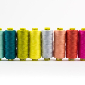 May include: Eight spools of sewing thread arranged in a row, featuring a gradient of colors including light blue, teal, lime green, bright yellow, silver, peach, orange-red, and magenta, each topped with a yellow label with black text.