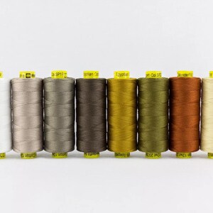 May include: Eight cylindrical spools of sewing thread arranged in a horizontal row. The colors range from white, light gray, medium gray, dark brown, mustard yellow, olive green, rust orange, to cream. Each spool has a bright yellow label.