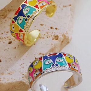 May include: Two open-band rings, one gold-toned and one silver-toned, featuring a colourful enamel pattern of abstract faces in blue, yellow, pink, and teal. Each ring includes a row of small, clear, sparkling stones set along the centre band.