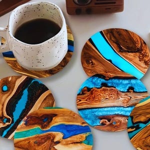 May include: A set of circular coasters made from natural wood and bright blue resin. Each coaster features a unique wood grain pattern with a smooth, glossy finish. One coaster supports a white speckled ceramic mug filled with dark liquid.