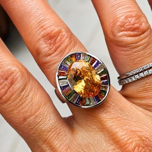 May include: A silver-toned ring featuring a large, oval-cut orange centre stone surrounded by a halo of multi-coloured, baguette-cut stones in shades of purple, green, orange, and white, set within a polished metal frame on a finger.