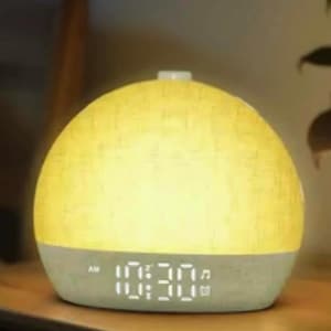 May include: A dome-shaped alarm clock with a textured yellow upper half and a solid light grey base. The digital display shows the time 10:30 AM in white light, accompanied by small musical note and calendar icons on the right side of the screen.