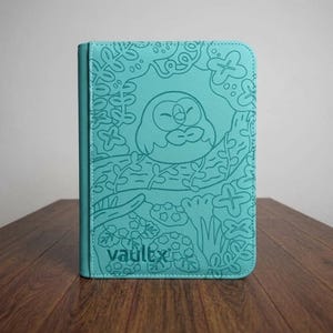 Può includere: Un raccoglitore rettangolare per carte collezionabili color verde acqua con un design a linee in rilievo che raffigura un uccello rotondo appollaiato su un ramo circondato da fogliame. Il marchio vaultx è impresso nell'angolo in basso.