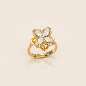 May include: A gold ring with a flower-shaped design. The flower has four white, oval-cut stones at the centre, encircled by smaller clear stones. The ring's band is adjustable.