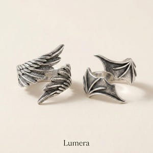 Puede incluir: Dos anillos de metal plateado con acabado oxidado. Un anillo presenta un diseño detallado de ala emplumada, mientras que el otro muestra una estructura de ala de murciélago. La palabra Lumera está impresa en negro en la parte inferior.