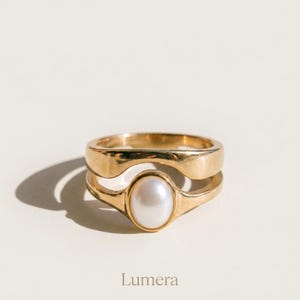 May include: A gold ring featuring a smooth, polished band with an open-work design and a single, centered oval white pearl. The word Lumera is printed in a simple, dark font below the ring, which is set against a plain, light-colored background.