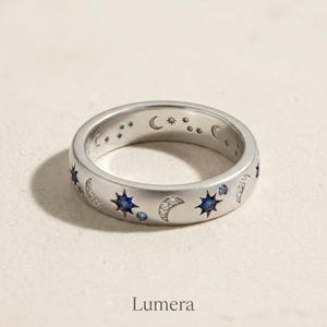 Può includere: Un anello d'argento con un design celeste. L'anello presenta pietre preziose blu a forma di stella, mezzelune e piccoli accenti di stelle. La parola "Lumera" è stampata in basso nell'immagine.