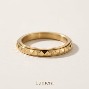 Op de afbeelding: Een goudkleurige metalen ring met een centrale band voorzien van een herhalend piramidevormig noppenpatroon. De ring heeft een glad binnenoppervlak en een gepolijste afwerking. Het woord Lumera staat in een donker lettertype onder de ring.