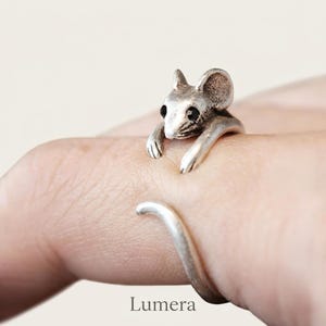 Op de afbeelding: Een zilverkleurige metalen ring in de vorm van een muis met zwarte kraaloogjes. De kop en voorpootjes van de muis rusten op de vinger, terwijl de lange, gebogen staart zich eromheen wikkelt en zo de verstelbare ringband vormt.