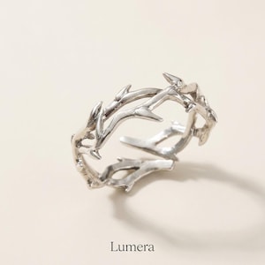 May include: A silver-toned ring featuring a textured design that mimics the appearance of intertwined thorny branches. The metal band has an open, adjustable structure with pointed, thorn-like protrusions extending along the entire circumference.