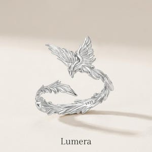 May include: A silver-colored open ring featuring a detailed bird design with wings spread and a long, curved tail. The band is textured with feather-like engravings and includes the stamp S925 on the inner surface. The word Lumera is below.