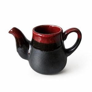 Op de afbeelding: Een keramische theepot met een tweekleurig glazuur, met een diepzwarte basis die overgaat in een felrode rand. De theepot heeft een gebogen tuit, een ronde vorm en een stevig handvat, afgewerkt met een gladde en glanzende bovenkant.