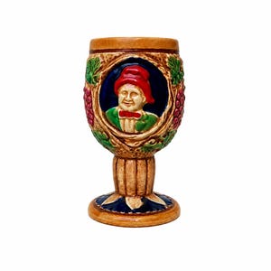 May include: A ceramic goblet featuring a relief portrait of a figure wearing a red hat and green jacket, set against a dark blue oval background. The exterior is decorated with sculpted grape clusters and leaves in shades of red and green.