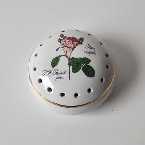 Könnte beinhalten: Ein weißes Potpourri-Gefäß aus Porzellan mit einem gewölbten Deckel, der am Rand mit einer Reihe kleiner kreisförmiger Löcher versehen ist. Der Deckel ist mit einer botanischen Illustration einer rosa Rosa centifolia und dem Text P.J. Redoute pinx. verziert.