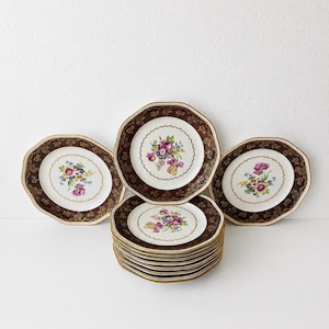 May include: A set of octagonal plates featuring a white center with a colorful floral bouquet, surrounded by a wide dark brown border with gold filigree patterns and a scalloped gold rim. The plates are arranged in a stack and fanned out.