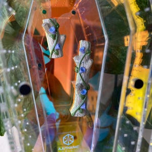 May include: Two miniature tree trunk figurines with blue flower vines inside a clear, geometric container. The container has a yellow and orange tint, with the word "Tarantula" visible. The figurines are light brown and gray.