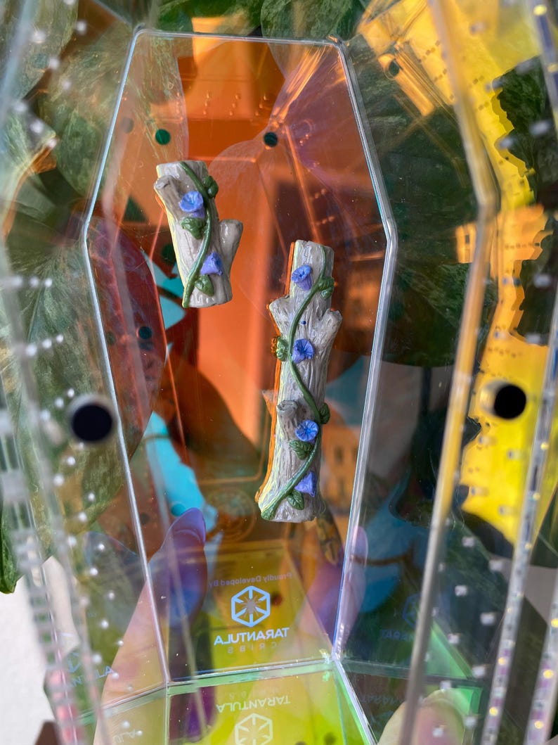 May include: Two miniature tree trunk figurines with blue flower vines inside a clear, geometric container. The container has a yellow and orange tint, with the word "Tarantula" visible. The figurines are light brown and gray.