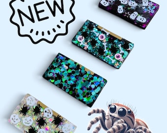 May include: Four rectangular resin dominoes featuring various Halloween-themed embeds including white ghosts, black spiders, jack-o'-lanterns, eyeballs, bones, and colorful glitter. A black outline graphic with the text NEW is in the top left corner.