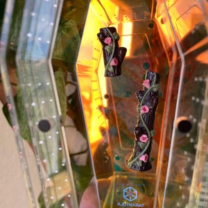 May include: Two miniature, dark brown tree trunk figurines with pink roses and green vines. The figurines are inside a clear, geometric container with the word "Tarantula" visible. The container has a yellow and orange tint.