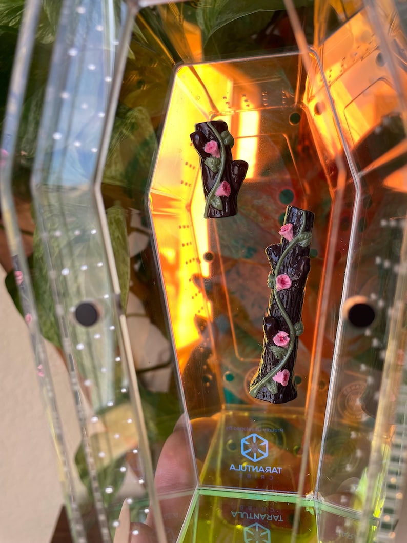 May include: Two miniature, dark brown tree trunk figurines with pink roses and green vines. The figurines are inside a clear, geometric container with the word "Tarantula" visible. The container has a yellow and orange tint.