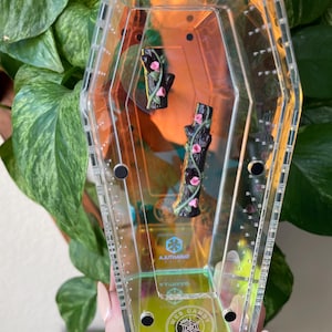 May include: A clear, coffin-shaped acrylic container with black accents and a yellow base. Inside are two miniature black tree trunk figurines adorned with pink flowers. The container has the text "WEB CANDY" and "AJUTHARAT".