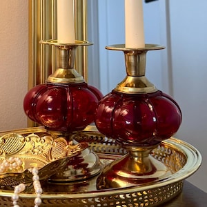 May include: Two brass candlesticks with red glass bases holding lit white taper candles, placed on a round gold-toned metal tray. A small gold-toned decorative dish containing a string of white faux pearls sits on the tray next to the candlesticks.