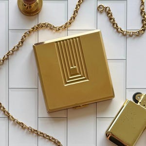 May include: A square gold-toned metal compact case featuring a centered geometric pattern of raised vertical lines and nested rectangles. The compact is surrounded by a delicate gold-toned chain and accompanied by a small gold-toned rectangular lighter.
