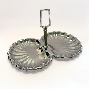 May include: A silver-toned metal serving dish featuring two connected shell-shaped bowls with scalloped edges. A central vertical handle with a rectangular loop at the top connects the two bowls, providing a structured design for serving.