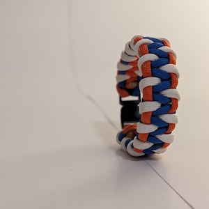 May include: A paracord bracelet featuring a braided pattern in white, orange, and blue nylon cord. The bracelet is secured with a black plastic side-release buckle, showcasing a thick, textured weave design that wraps around the wrist.