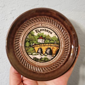 May include: A round ceramic coaster with a dark brown glazed rim featuring a textured rope-like pattern. The center displays a colorful illustration of a stone bridge over water with a building and trees, with the word Randaya printed above.