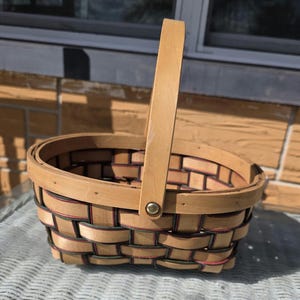 May include: An oval-shaped woven basket constructed from light-colored wooden splints. The basket features a single upright handle attached with a metal rivet and is accented with thin, dark-colored horizontal bands woven throughout the body.