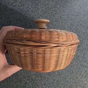 May include: A round, light brown woven wicker basket with a matching lid. The lid features a centered, smooth, light-colored wooden knob handle. The basket has a horizontal weave pattern with vertical structural supports visible around the exterior.