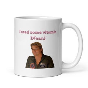 Puede incluir: Una taza de cerámica blanca con la imagen impresa de una persona de cabello castaño claro que viste una chaqueta de vuelo verde oscuro con parches. Sobre la imagen, el texto "I need some vitamin D(ean)" aparece en letras rosas.
