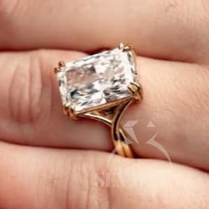 Peut inclure: Une bague en or ornée d'une grande pierre transparente rectangulaire à taille rayonnante, maintenue par quatre griffes. L'anneau est fin et lisse, se connectant au sertissage avec un design effilé. La pierre est facettée et reflète la lumière.
