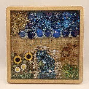 May include: A square wooden shadow box frame containing a mixed media collage. The top section features gold leaf and blue glass gems. The bottom section displays sunflowers, daisies, a small turtle figurine, blue stones, and dried foliage.