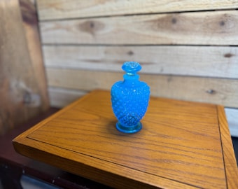 Puede incluir: Un pequeño frasco de perfume de vidrio azul brillante con un patrón texturizado de puntos en su cuerpo redondeado. El frasco incluye un tapón de vidrio azul a juego que encaja en el cuello estrecho y descansa sobre una base circular.