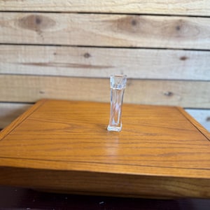 Puede incluir: Un pequeño frasco de perfume de cristal transparente con base rectangular y cuerpo esbelto ligeramente cónico. El frasco es transparente y presenta un diseño liso y sencillo con un centro hueco y una abertura superior plana.