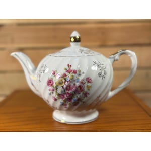 Puede incluir: Una tetera de porcelana blanca con una textura acanalada en espiral y un ramo floral central de flores rosas, amarillas y azules. La tetera incluye una tapa a juego con un remate dorado, un asa curva y un pico con detalles en color oro.