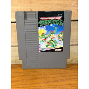 Pode incluir: Um cartucho de videogame cinza para Nintendo Entertainment System de Teenage Mutant Ninja Turtles. O rótulo apresenta os personagens de tartarugas verdes em poses de ação com máscaras vermelhas, o título do jogo em letras verdes e o logo Ultra.