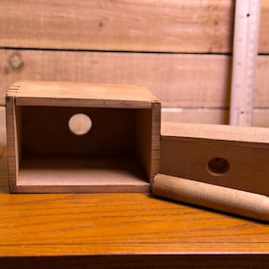 Könnte beinhalten: Eine hellbraune Holzschachtel mit gezinkten Ecken und einem kreisförmigen Loch an der Vorderseite. Neben der Schachtel liegt ein passender Holzdeckel mit einer kreisförmigen Vertiefung sowie ein zylindrischer Holzstab, alles mit Lineal.