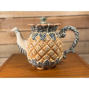 May include: A ceramic teapot featuring a textured pineapple-inspired body with a golden-yellow diamond pattern and dark blue leaf accents. The lid has a matching leaf design, and the handle is shaped with a twisted, rope-like appearance.