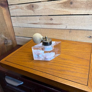 Pode incluir: Um frasco de perfume de vidro transparente com base hexagonal e um design floral interno. O frasco é rematado com um colar metálico e uma bomba atomizadora de pera texturizada na cor creme, um acessório de toucador vintage.
